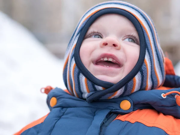 Attraente bambino che gioca con la prima neve. Sorride e sembra pupazzo di neve. Tuta spessa blu-arancio cappello a righe luminose su un bambino di anno . Foto Stock Royalty Free