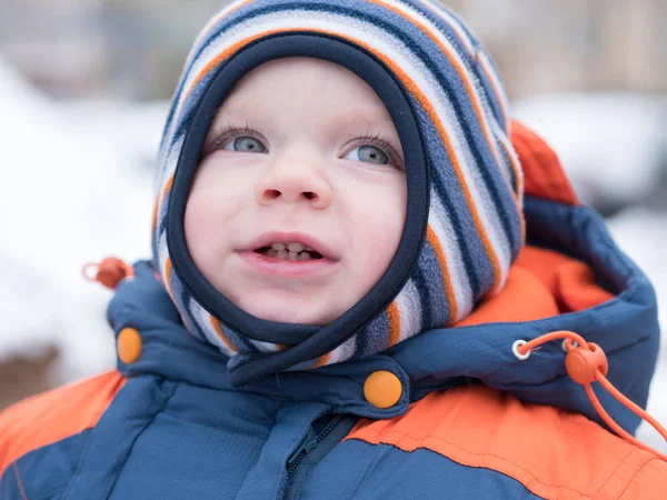 Menino atraente brincando com a primeira neve. Ele sorri e parece boneco de neve. Macacão azul-laranja grosso chapéu listrado brilhante em uma criança de ano . Imagem De Stock