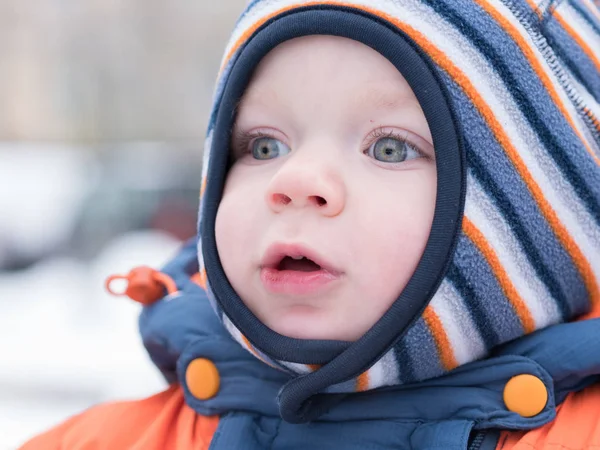 Menino atraente brincando com a primeira neve. Ele sorri e parece boneco de neve. Macacão azul-laranja grosso chapéu listrado brilhante em uma criança de ano . Imagem De Stock