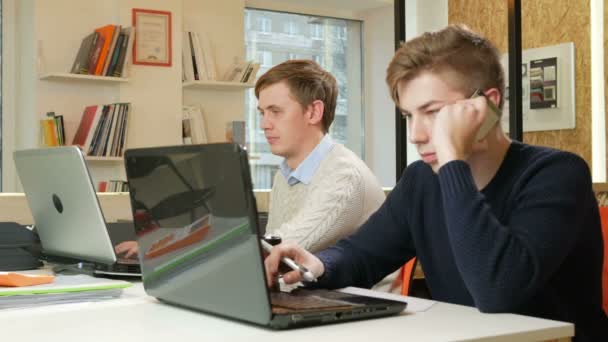 Молодой человек работает в офисе ноутбука над проектом. Один из них звонит по телефону клиенту и записывает ответы. Концепция командной работы — стоковое видео
