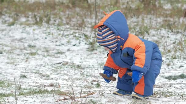 Aantrekkelijke baby spelen in het bos van de winter met haar moeder. Op de grond, een beetje sneeuw. Jongen spelen met sabels en takken. De warme blauwe en oranje jumpsuit — Stockvideo