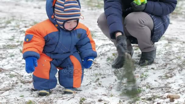 魅力的な赤ちゃんが母親と冬の森で遊ぶ。地上では、雪のビット。サーベルと枝で遊ぶ少年。温かみのあるブルーとオレンジのジャンプ スーツ — ストック動画