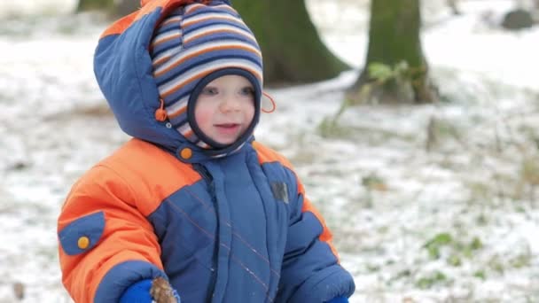 Aantrekkelijke baby spelen in het bos van de winter met haar moeder. Op de grond, een beetje sneeuw. Jongen spelen met sabels en takken. De warme blauwe en oranje jumpsuit — Stockvideo