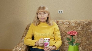 Kadın evde kanepede bir fincan sıcak bir içecek içme ve kameraya bakarak yaşında. Zor bir günün ardından dinleniyor. tatil kavramı