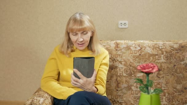 Seniorin mit Tablet sitzt auf Couch im Wohnzimmer. spricht Videokommunikation mit Verwandten. er lächelt und spricht über sich selbst — Stockvideo