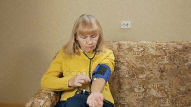 Yaşlı kadın kendi evinde kanepede basınç ölçer. O darbe için Tansiyon aleti ile dinler