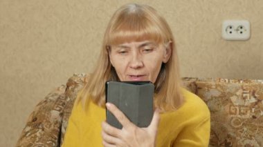 Oturma odasında kanepede oturan tablet ile kadın kıdemli. Video iletişim akrabaları ile konuşuyor. O gülümseyen ve kendisi hakkında konuşmak