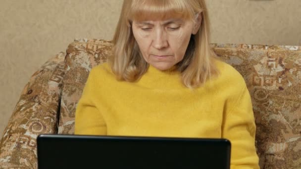 Mulher envelhecida trabalha cuidadosamente em um laptop em casa no sofá. Ela está digitando no teclado e olhando para a tela. O conceito de trabalhar em casa — Vídeo de Stock