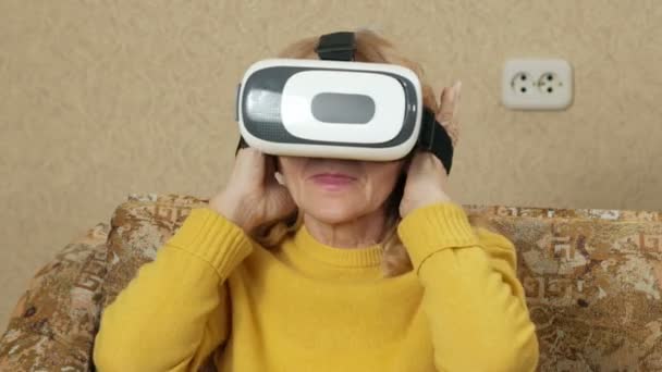 Une femme âgée porte des lunettes de réalité virtuelle et regarde un film. Elle regarde autour d'elle et veut toucher les murs virtuels. concept de maison de vacances — Video