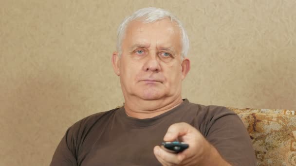 Мужчина в возрасте переключает телеканалы дистанционного управления на диване дома. Он расслабляется после тяжелого дня. Концепция дома отдыха — стоковое видео