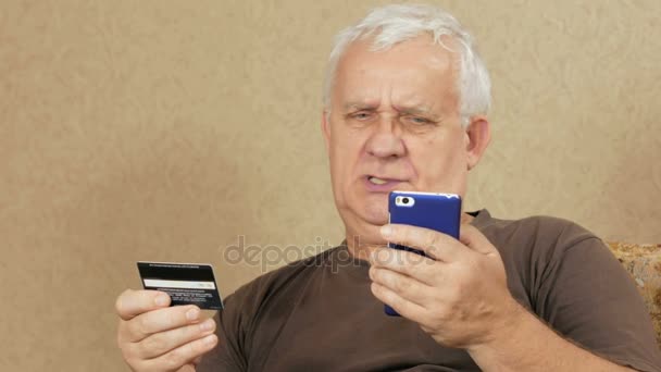 Старший чоловік оплачувати покупки в Інтернет-банк кредитну картку. Він уважно представляє номер кредитної картки на телефон. концепції home shopping — стокове відео