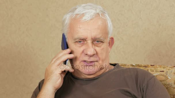 Mężczyzna w wieku rozmawia przez telefon komórkowy w domu na kanapie. Wyraz jego twarzy zmienił. komunikacja koncepcja — Wideo stockowe