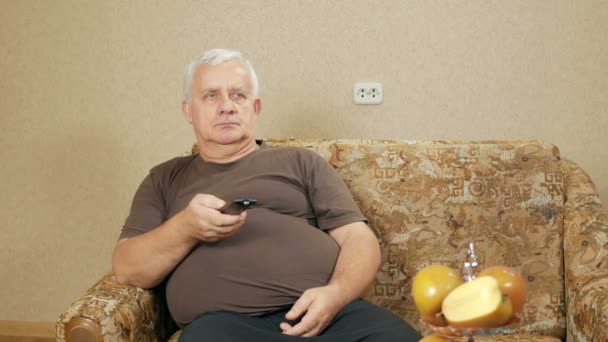 Man aged switches remote control TV channels on the couch at home. He relaxes after a hard day. holiday home concept — Stock Video