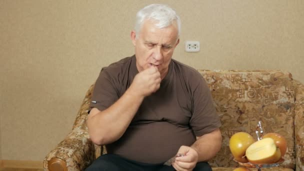 Hombre mayor enfermo con sobrepeso tomando pastillas en casa en un sofá y bebe agua en un vaso. Hace varios líquidos suaves. El concepto de salud en los ancianos — Vídeos de Stock