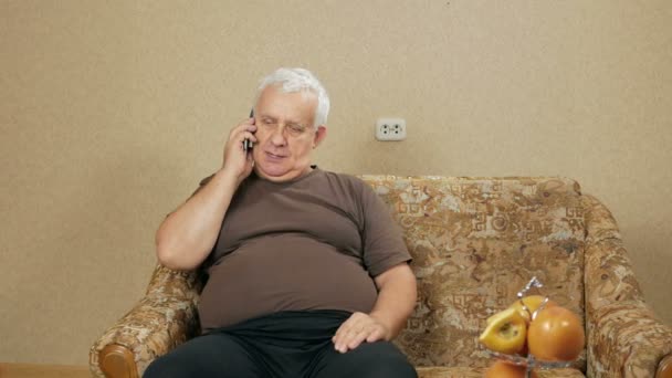 Мужчина в возрасте разговаривает по сотовому телефону дома на диване. Его выражение лица изменилось. коммуникационная концепция — стоковое видео