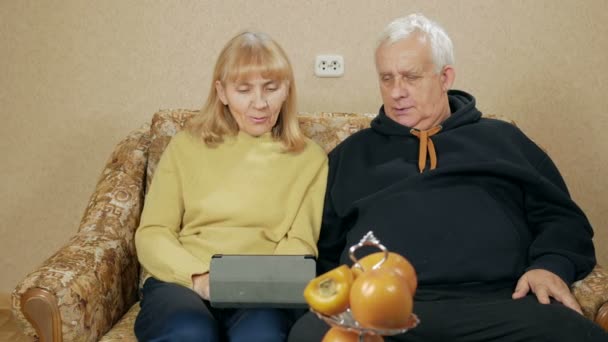 Пожилая пара звонит по видеосвязи на планшете. Муж и жена сидят дома на диване. Концепция общения близких родственников — стоковое видео