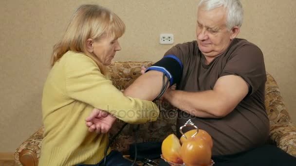 年配の女性は、ソファの上の男は自宅の血圧を測定します。彼女はあなたの腕にカフを着ているし、パルス血圧計に耳を傾けます。健康の概念 — ストック動画