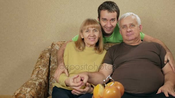 Joven hijo de una pareja de ancianos abrazándose en casa en el sofá. Todos miran a la cámara y sonríen. Se comunican entre sí y se ríen. concepto familiar feliz . — Vídeos de Stock