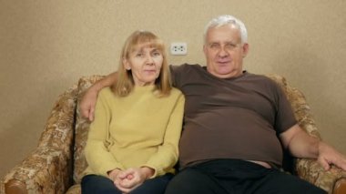 Yaşlı çift, evde koltukta istirahat ve kameraya bakıyor. Onlar gülüyor. Kocası karısını sarılmak.