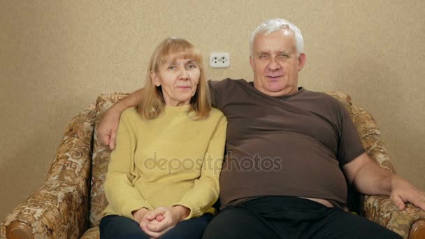 Elderly couple resting at home on the couch and looking at the camera. They are smiling. The husband hug his wife. — Stock Video