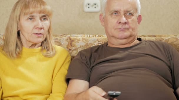 一对老年夫妇在家里看电视在沙发上。女人笑着拿电视遥控器本身和选择有趣的节目。假期的家居理念 — 图库视频影像