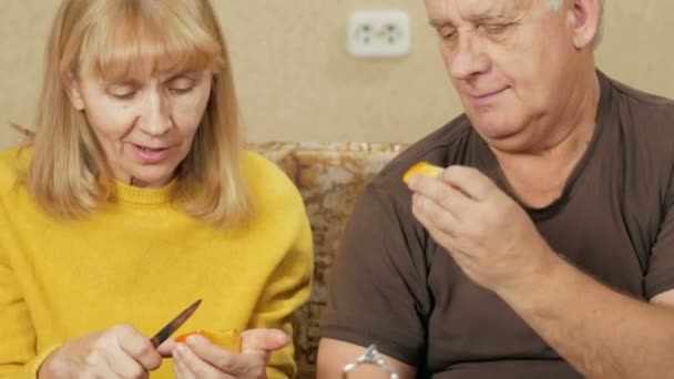 年长的夫妇在沙发上吃水果在家里。女子切下一块柿子，给男性超重。健康食品的概念 — 图库视频影像