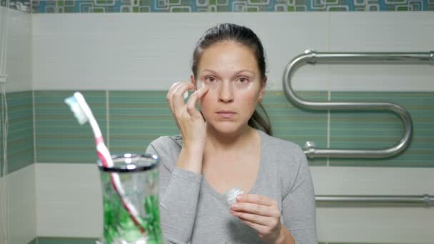 Giovane donna attraente applica crema per la pelle intorno agli occhi in bagno. Lei fa un punto della crema e poi strofinando delicatamente le dita — Video Stock