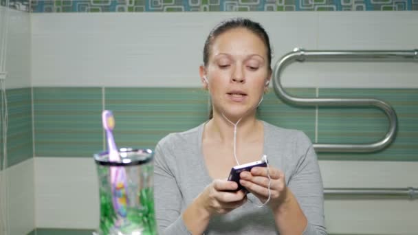 Молода приваблива жінка слухає музику з навушниками по телефону у ванній кімнаті готелю. Вона посміхається до камери і співає улюблену пісню — стокове відео