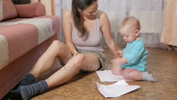 Criança atraente desenha um lápis no chão com a mãe em casa. O rapaz está a sorrir e a brincar com a mãe. O conceito de férias em família — Vídeo de Stock