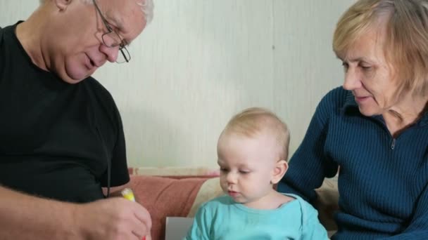 Menino atraente desenha uma caneta com seus avós em casa no sofá. O menino olha para os animais que atraíram o avô. O conceito de diferentes gerações — Vídeo de Stock