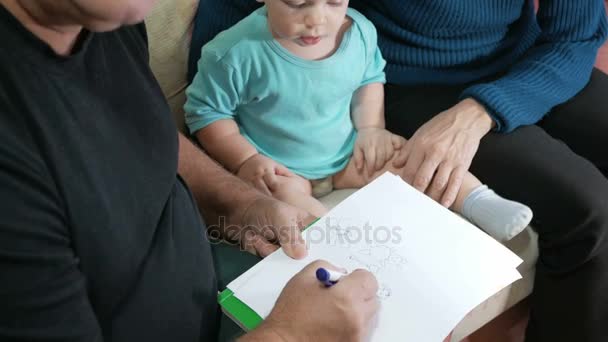 Attractive baby boy draws a pen with his grandparents home on the couch. The boy stares at the animals that drew grandfather. The concept of different generations — Stock Video
