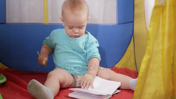 Attraktiva baby spelar i barnens färgglada tältet i huset. Pojken drar försiktigt i penna anteckningsbok. — Stockvideo