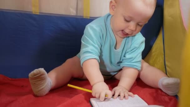 Atractivo bebé jugando en la carpa colorida de los niños en la casa. El niño dibuja cuidadosamente en cuaderno de lápiz . — Vídeos de Stock