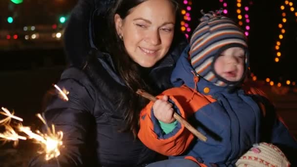 La madre joven con un niño celebra el año nuevo en la calle con un bengala. Abrazando y sonriendo a la cámara. En el fondo está decorado árbol de Navidad — Vídeo de stock