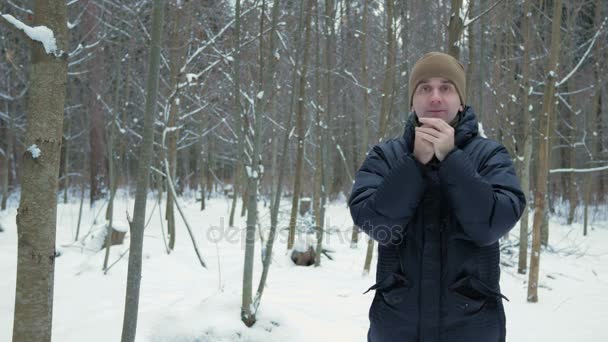一个年轻人在冬季森林冻结。他在他的手，呼吸声揉和戴帽夹克。雪域景观。他看着镜头 — 图库视频影像