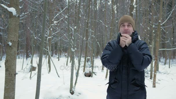 Un joven congelado en el bosque de invierno. Respira en sus manos, frota y usa chaquetas de capucha. Paisaje nevado. Mira a la cámara. — Vídeos de Stock