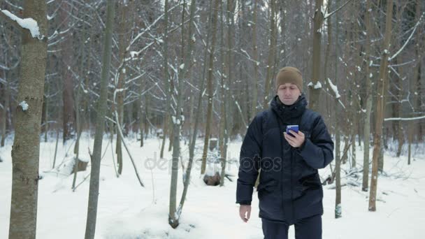 Le jeune homme va dans la forêt d'hiver et est orienté au téléphone. Il cherche la bonne direction sur la carte électronique. Un homme vêtu d'une veste sombre et d'un chapeau chaud . — Video