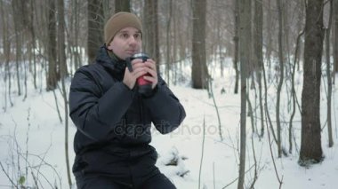 Bir kış ormandaki bir termos sıcak bir içecek içme bir genç adam. Adam kar ve ağaçlar bir koyu renk ceketli ve sıcak bir şapka etrafına bakıyor. doğa gevşeme kavram