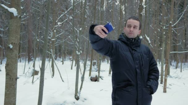 Homem faz telefone selfie em uma floresta de inverno nevado. Ele sorri e muda a postura para uma foto melhor. disponibilidade conceito foto — Vídeo de Stock