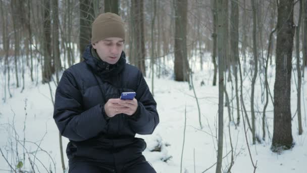 Young man typing a sms message on the phone in the winter snowy forest. He smiles and speaks the written. — Stock Video