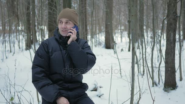 Young man sitting in the winter forest and talking on the phone. He admires the sides of snow and trees. A man in a dark jacket and a warm hat. — Stock Video