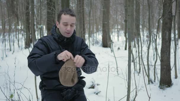 Молодой человек в зимнем лесу замерз. Он дышит на руках, массирует и носит шляпу и перчатки. Снежный пейзаж . — стоковое видео
