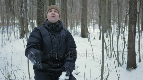 Il giovane guarda nella macchina fotografica e sorride nella foresta invernale. Giocare con la neve. Un uomo con una giacca scura e un cappello caldo . — Video Stock