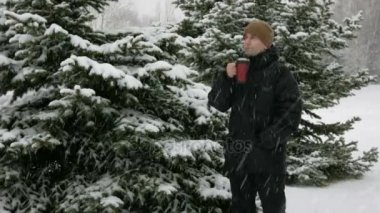 Bir kış ormandaki bir termos sıcak bir içecek içme bir genç adam. Büyük kar yağışı. Adam kar ve ağaçlar bir koyu renk ceketli ve sıcak bir şapka etrafına bakıyor. doğa gevşeme kavram