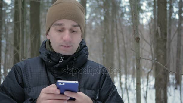 Mladý muž píše zprávu sms na telefonu v zimě zasněženém lese. Usměje se a hovoří písemné. — Stock video