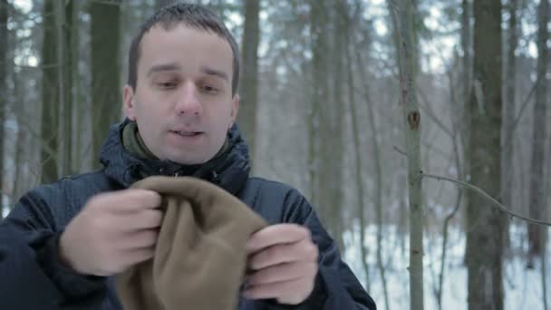 A young man in winter forest freeze. Hes breathing on his hands, rubs and wears a hat and gloves. Snowy landscape. He looks into the camera — Stock Video