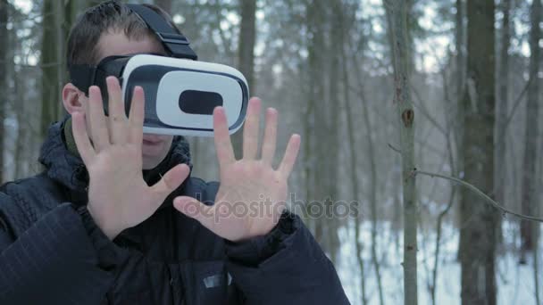 En man klädd i en virtuell verklighet headset på vintern i en snöig skog. Han försöker röra virtuella objekt. En man klädd i en varm jacka och vifta med armarna. — Stockvideo