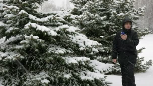 De jonge man gaat in een bos van de winter in de sneeuw en is gericht op de telefoon. Hij is op zoek naar de juiste richting op de elektronische kaart. Een man in een donkere jas en warme capuchon. — Stockvideo