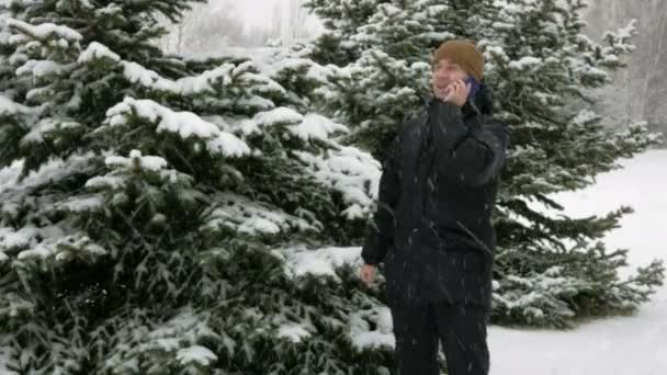Um jovem vai para uma floresta de inverno e fala ao telefone. Grande queda de neve. Ele pára e termina a conversa. Um homem com um casaco escuro e um chapéu quente . — Vídeo de Stock
