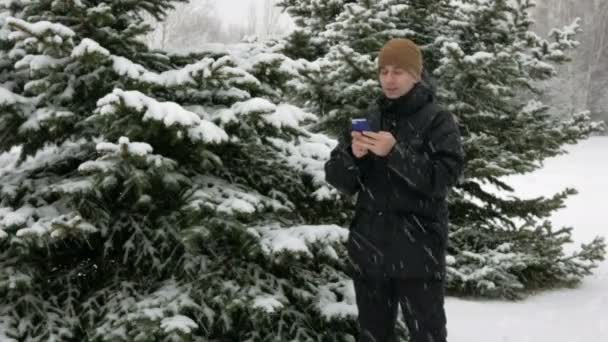 Homem jovem digitando uma mensagem sms no telefone na floresta nevada de inverno. Grande queda de neve. Ele sorri e fala o escrito . — Vídeo de Stock